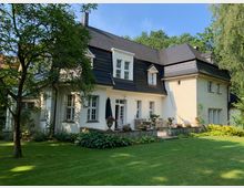 Zweistöckiges Einfamilienhaus mit hellem Putz, dunklem Walmdach und mehreren weißen Sprossenfenstern, umgeben von einem gepflegten Garten mit Bäumen, Rasen und einer Terrasse mit Sitzmöbeln.