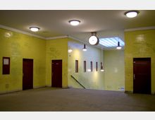 U-Bahn-Station mit gelben Wandfliesen, roten Türen und kleinen, rechteckigen Fenstern. Von der Decke hängen Lampen, und eine große Uhr ist zentral angebracht; eine Treppe führt nach unten.