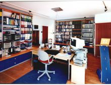 Ein gut organisiertes Büro mit Holzfußboden, großen Regalen voller Aktenordner und Büchern, einem Schreibtisch mit Computer und Bürozubehör sowie einer Schreibtischlampe; rechts im Bild steht ein roter Heizkörper und ein moderner Prospektständer.