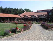 Hof mit gepflastertem Weg, der zu einem Gebäudeensemble mit roten Ziegeldächern und Holzfassaden führt, umgeben von Blumenbeeten mit Rosen und Lavendel. Im Hintergrund sind Bäume eines Waldes zu sehen.