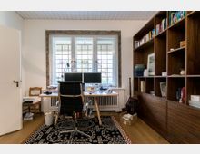 Helles Arbeitszimmer mit großer Fensterfront und Gitterverglasung, einem Schreibtisch mit Doppelmonitoren und einem schwarzen Bürostuhl. Rechts befinden sich deckenhohe Holzbücherregale, links ein offener Türbereich und ein Stuhl, auf dem Boden liegt ein gemusteter Teppich.