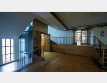 Flur eines Gebäudes mit Treppenhaus, einem geöffneten Aufzug mit Holztür und einer Wendeltreppe mit Metallgeländer. Große Glasfenster lassen Tageslicht herein, und eine Notausgangstür sowie ein Feuerlöscher sind sichtbar.