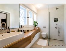 Modernes Badezimmer mit einem großen Waschtisch aus Holz und einem weißen Waschbecken. Rechts befindet sich eine bodenebene Dusche mit Glasabtrennung und ein wandmontiertes WC; die Fenster mit Gittereinsätzen lassen Tageslicht herein.