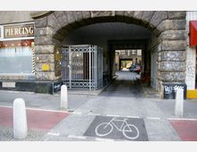 Steinernes Torbogen-Gebäude mit angrenzendem Schaufenster eines Piercingstudios auf der linken Seite und einem verschlossenen Metallgitter vor dem Durchgang. Im Hintergrund ist ein Innenhof mit geparkten Autos sichtbar, während Pflastersteine und ein Fahrradweg im Vordergrund den Boden markieren.