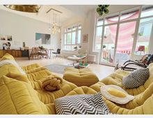 Helles Wohnzimmer mit großen Fenstern und Zugang zu einem Balkon. Im Vordergrund ein gelbes Sofaset mit dekorativen Kissen, im Hintergrund ein Esstisch und moderne Einrichtungselemente.