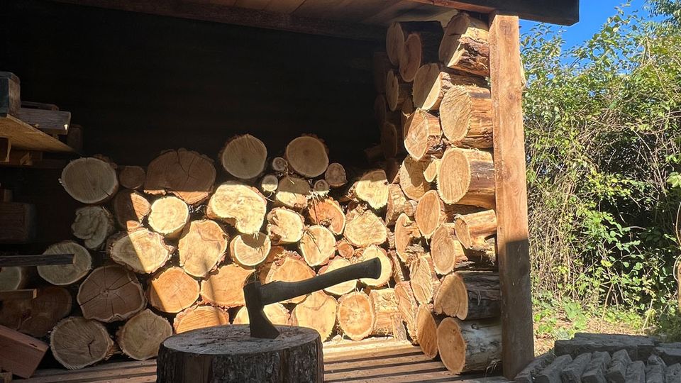 Offener Holzunterstand im Freien, gefüllt mit gestapelten Baumstämmen und Rundhölzern unter einem flachen Dach. Davor steht ein Hackklotz mit einem Beil; umgeben von Kiesboden, Büschen und Bäumen unter blauem Himmel.