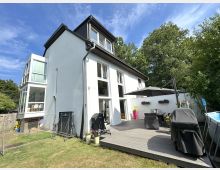 Modernes, weiß verputztes Einfamilienhaus mit dunklem Satteldach, großen Fensterflächen und einem gläsernen Anbau an der Seite. Davor liegt eine Holzterrasse mit Gartenmöbeln, Sonnenschirm und Grill, umgeben von Rasen und dichtem Baumbestand.