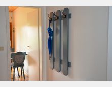 Flur mit einer schlichten Garderobe aus Holzlatten, an der ein blauer Regenschirm hängt. Im Hintergrund ist durch eine geöffnete Tür ein Essbereich mit Holzstühlen und gepolsterten Rückenlehnen sichtbar.