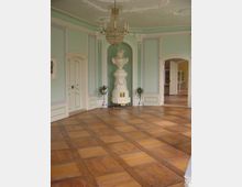 Eleganter historischer Raum mit hellgrünen Wänden, weißen Zierleisten und hohen Türen. Ein zentraler Kachelofen in hellem Weiß, eine große Kristall-Deckenleuchte und ein gealterter Holzfußboden prägen das Ambiente.