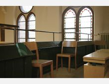 Innenraum einer Kirche mit Holzbänken und Stühlen, ergänzt durch farbige Glasfenster in gotischem Stil mit roten und grünen Mustern. Im Hintergrund ist ein Heizkörper an der Wand sichtbar.