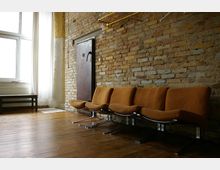 Raum mit einer Wand aus freiliegenden Ziegeln, einer Sitzreihe mit gepolsterten, orangefarbenen Stühlen auf Metallgestellen und einem Holzboden. Im Hintergrund befindet sich ein großes Fenster mit weißen Vorhängen und ein dunkler Türrahmen.