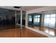 Ein Trainingsraum mit hellem Laminatboden, großen Wandspiegeln und bodenlangen Fenstern mit blauen Vorhängen. In der Mitte des Raums befinden sich mehrere vertikale Metallstangen, die offenbar für Pole-Fitness oder Tänze genutzt werden.