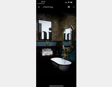 Ein Badezimmer mit dunklem Ziegelmauerwerk und einer freistehenden weißen Badewanne mit goldfarbener Armatur. An der Wand hängt ein Schwarz-Weiß-Druck mit dem Wort "hi", daneben ein sechseckiger Spiegel; im Hintergrund befinden sich Regale mit gerollten Handtüchern.