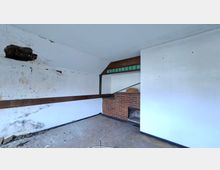 Innenraum eines leerstehenden, stark beschädigten Zimmers mit abblätterndem Putz, Rissen in der Decke und einem großen Loch in der linken Wand. Rechts steht eine gemauerte Ziegelkonstruktion mit offenem Kamin oder Nische, darüber ein Holzaufsatz mit schmalen grünen Glasfenstern.
