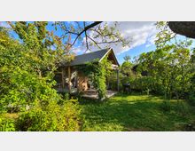 Kleines Holzhaus mit großen Glasfenstern und einer überdachten Terrasse, umgeben von dichtem, grünen Garten mit Bäumen, Sträuchern und einer Rasenfläche. Im Vordergrund ist ein Grill erkennbar, bei hellem Tageslicht und blauem Himmel.