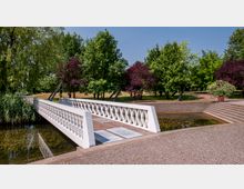 Weiße Fußgängerbrücke mit durchbrochenem Geländer, die über ein flaches Gewässer führt, umgeben von Bäumen und Grünflächen in einem Park; im Hintergrund eine gepflasterte Terrasse mit Stufen.