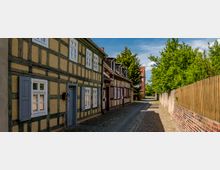 Schmale gepflasterte Gasse mit Fachwerkhäusern auf der linken Seite, gekennzeichnet durch gelbe und braune Fassaden sowie blaue Fensterläden. Auf der rechten Seite stehen eine Holzlatten- und eine Backsteinmauer, flankiert von Bäumen und Sträuchern.