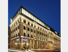 Ein großes, mehrstöckiges Gebäude im neoklassischen Stil, beleuchtet bei Nacht mit einer Fassade aus steinernen Bögen und Fenstern; auf dem Dach befindet sich ein modernes, verglastes Aufbauelement. Im Vordergrund sind Straßen mit Lichtspuren von fahrenden Autos zu sehen.