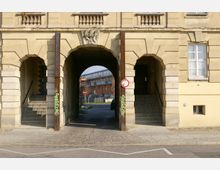 Sandfarbene Fassade eines historischen Gebäudes mit drei bogenförmigen Durchgängen; der mittlere führt auf eine Straße und zeigt im Hintergrund moderne Wohngebäude. Die Seitendurchgänge enthalten Treppenaufgänge, und die mittlere Arkade ist mit Ornamenten und einem Wappen geschmückt.
