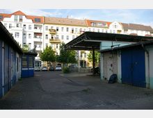 Hinterhof mit gepflastertem Boden, umgeben von Gebäuden mit blauen Türen und Garagen auf der linken und rechten Seite. Im Hintergrund mehrstöckige Wohnhäuser mit weißen Fassaden, roten Dächern und einigen Balkonen, sowie parkende Autos und Bäume.