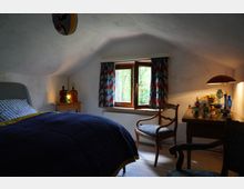 Ein kleines Schlafzimmer mit einem schrägen Dach, einem Holzbett mit dunkler Bettdecke, einem Fenster mit bunten Vorhängen und einem Nachttisch mit Glasflaschen. Ein Holztisch mit dekorativen Glasobjekten und eine Lampe stehen neben einem Stuhl aus Holz.