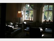 Wohnzimmer mit großen, weißen Sprossenfenstern, durch die Tageslicht hereinfällt. Der Raum ist mit einem grauen Ledersofa, einem Beistelltisch und einem Stehlampenlicht ausgestattet; im Fensterbereich stehen dekorative Glasgefäße und kleine Pflanzen.