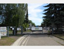 Eingangstor eines Betriebsgeländes mit Gitterzaun, Hinweisschildern und einem Banner mit der Aufschrift „Demontage Schulz“. Im Hintergrund sind Bäume, Fahrzeuge und Container zu sehen.