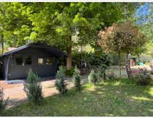 Kleines, dunkles Holzhaus mit Vordach inmitten eines Gartens, umgeben von hohen Bäumen, Sträuchern und gepflegtem Rasen. Im Vordergrund stehen junge Büsche und ein kleiner Baum mit rötlichem Laub.