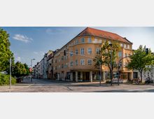 Eckhaus in einem Stadtviertel mit gelbem Putz und rotem Walmdach, großen rechteckigen Fenstern und einem überdachten Eingangsbereich. Im Vordergrund eine Kreuzung mit Ampeln, Bäumen und vereinzelt parkenden Fahrrädern.