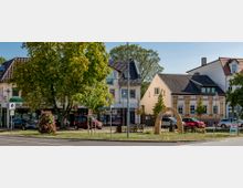 Städtische Szene mit mehreren zweigeschossigen Gebäuden, darunter Geschäfte mit Schaufenstern im Erdgeschoss. Im Vordergrund ein kleiner Platz mit Bäumen, Blumenbeet, Sitzbänken und einem dekorativen Steinbogen.