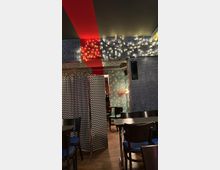 Ein stilvoll eingerichtetes Café mit dunklen Holztischen und schwarzen Stühlen mit blauen Sitzpolstern. Die Wände sind mit gemusterten Tapeten dekoriert, beleuchtet durch Lichterketten, und im Vordergrund steht ein Paravent mit Zickzack-Muster.