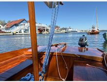 Blick vom hölzernen Deck eines kleinen Segelbootes über einen Hafenkanal, mit sichtbaren Leinen und Mast im Vordergrund. Am Ufer stehen Gebäude mit roten Dächern und Steganlagen, im Wasser liegen weitere Boote vor blauem Himmel.
