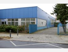 Flaches, modernes Gebäude mit blauer Metallfassade und großen Fenstern. Vor dem Gebäude liegt ein Gehweg mit Pflastersteinen, ein offenes blaues Tor sowie einige Büsche und ein Baum.