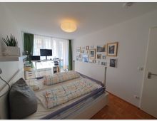 Schlafzimmer mit einem Doppelbett im Vordergrund, hellem Holzfußboden und weiß gestrichenen Wänden. An der rechten Wand hängt eine Galerie aus gerahmten Bildern, während ein Fenster mit grünen Vorhängen und einem kleinen Schreibtischbereich mit Monitor und Regal im Hintergrund zu sehen ist.