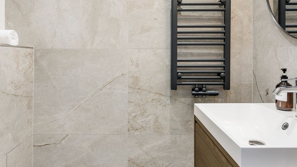 Modernes Badezimmer mit großformatigen, hellgrau marmorierten Wand- und Bodenfliesen. Zu sehen sind ein wandhängendes WC, ein Waschtisch mit Holzunterschrank, ein schwarzer Handtuchheizkörper sowie ein runder Spiegel und eine lineare Deckenleuchte.
