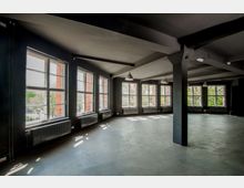 Großer, leerer Raum mit Betonboden, dunklen Wänden und zahlreichen großen Fenstern, die viel Tageslicht hineinlassen. An den Wänden sind schwarze Heizkörper und an der Decke hängen schlichte industrielle Lampen.