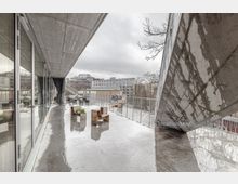 Moderne Terrasse mit Betonarchitektur, großflächigen Glasfenstern und einer Überdachung aus schrägen Betonplatten. Der Boden ist nass, es gibt einfache Holzbänke, Pflanzen in Kästen und einen Blick auf umliegende Gebäude und Bäume.