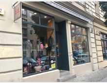 Straßenansicht eines Geschäftes mit der Aufschrift „designer depot“ oberhalb der großen Schaufenster. In den Fenstern sind Kleidungsstücke und Accessoires ausgestellt, während sich die Gebäudefassade durch beigen Putz und traditionelle Elemente auszeichnet.