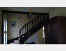 Innenraum mit einer hölzernen, geschwungenen Treppe, die zu einer oberen Etage führt. Der Raum hat helle Wände, ein großes Holzschrankmöbel rechts und Kunstwerke sowie Fenster an den linken Wänden.