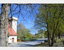 Eine Landschaft mit einem historischen Gebäude mit rotem Ziegeldach und einem weißen Turm im Vordergrund, umgeben von Bäumen mit frischem Frühlingslaub. Neben dem Gebäude stehen Holzbänke, und eine gepflasterte Straße führt durch die grüne Umgebung.