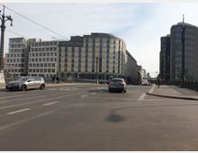 Straßenszene einer Brücke in einer Stadt mit modernen Bürogebäuden im Hintergrund, darunter ein oval geformtes Gebäude mit der Aufschrift "ME". Autos fahren und parken auf der Straße, während Passanten den Bürgersteig neben der Brücke benutzen.
