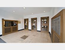 Eingangsbereich eines öffentlichen Gebäudes mit weiß gestrichenen Wänden und gefliesten Böden. Links befindet sich ein hölzerner Schalter mit Glasfenster, rechts eine Tafel mit der Aufschrift „Familiengericht“, und drei Holztüren mit Glaseinsätzen führen nach innen.