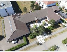 Luftaufnahme eines einstöckigen Gebäudekomplexes mit braunen Ziegeldächern und hellem Innenhof in rechteckigem Muster. Umgeben von Gehwegen, Grünflächen und benachbarten Wohngebäuden in einer ruhigen Wohngegend.