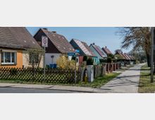 Straßenszene mit einer Reihe von Einfamilienhäusern mit Satteldächern und unterschiedlichen Fassadenfarben, entlang eines gepflasterten Gehwegs. Vorgärten mit Zäunen und Bäumen sowie ein Schild mit der Aufschrift "30-Zone" und ein Straßenschild sind zu sehen.