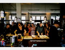 Eine Szene in einem modernen Restaurant oder Café mit großen Fenstern im Hintergrund. Menschen stehen an einer Theke, wo Essen zubereitet und serviert wird; im Vordergrund sind Küchenutensilien und Lebensmittel zu sehen.