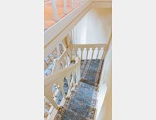 Treppenhaus mit weißem Geländer im neogotischen Stil, verziert mit Spitzbogenmotiven. Der Boden ist mit einem blauen Teppich mit floralem Muster bedeckt, darunter ist ein Raum mit Holzboden erkennbar.