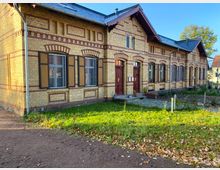 Historisches Wohnhaus aus gelbem Backstein mit dunkelroten Zierelementen und Holzfensterläden, flankiert von kleinen Vorgärten und umgeben von herbstlicher Vegetation. Das Gebäude hat ein spitzes Dach und mehrere Eingänge mit braunen Türen.