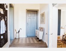 Flur in einem Wohnhaus mit einer hellblauen Tür, einem historischen, gemusterten Bodenfliesenbelag und zwei weißen Kinosesseln an der Wand. Neben der Tür steht ein Metallkrug, der als Schirmständer dient, und links an der Wand hängt eine schwarze Pinnwand.