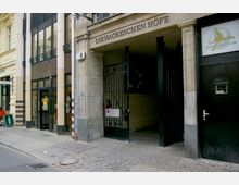 Eingang zu den Hackeschen Höfen in Berlin mit einer offenen schwarzen Eisentür. Daneben befinden sich Schaufenster mit Schaufensterpuppen sowie Gebäudedetails wie helle Fassaden und ein Pflasterstein-Bürgersteig.