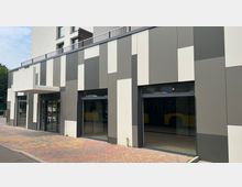 Fassade eines modernen Gebäudes mit einer Kombination aus grauen und weißen vertikalen Paneelen. Große Fensterfronten und ein kleiner, überdachter Eingangsbereich; gepflasterter Gehweg im Vordergrund.